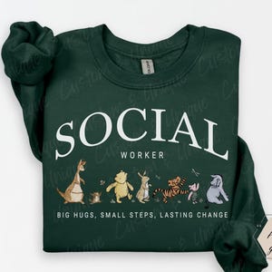 Pu&ograve; includere: Felpa verde scuro con la scritta bianca "SOCIAL WORKER" e una fila di personaggi dei cartoni animati. Sotto c'&egrave; la frase "BIG HUGS, SMALL STEPS, LASTING CHANGE".