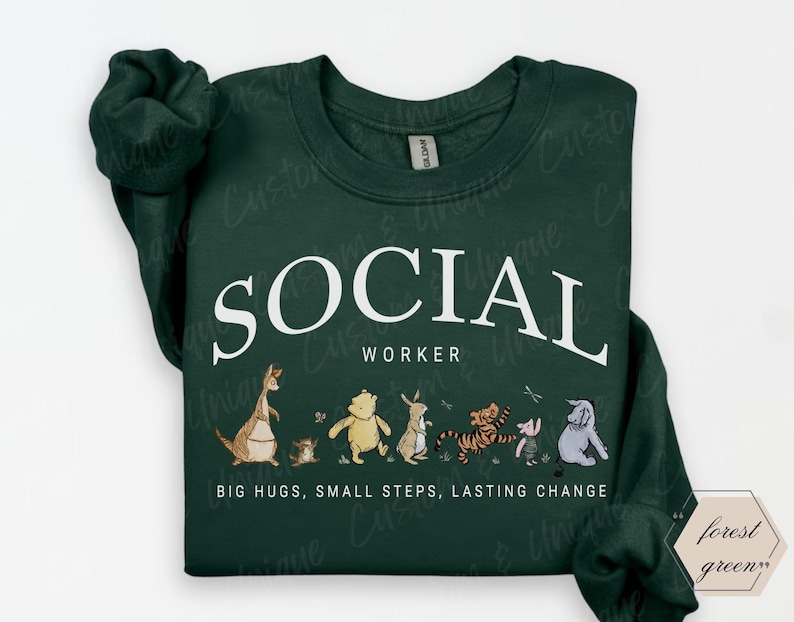 Pu&ograve; includere: Felpa verde scuro con la scritta bianca "SOCIAL WORKER" e una fila di personaggi dei cartoni animati. Sotto c'&egrave; la frase "BIG HUGS, SMALL STEPS, LASTING CHANGE".