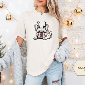 May include: Ivory-colored t-shirt featuring a graphic of a French Bulldog with reindeer antlers and a red nose, wrapped in Christmas lights. The dog is lying down. The shirt is part of a holiday-themed apparel collection.