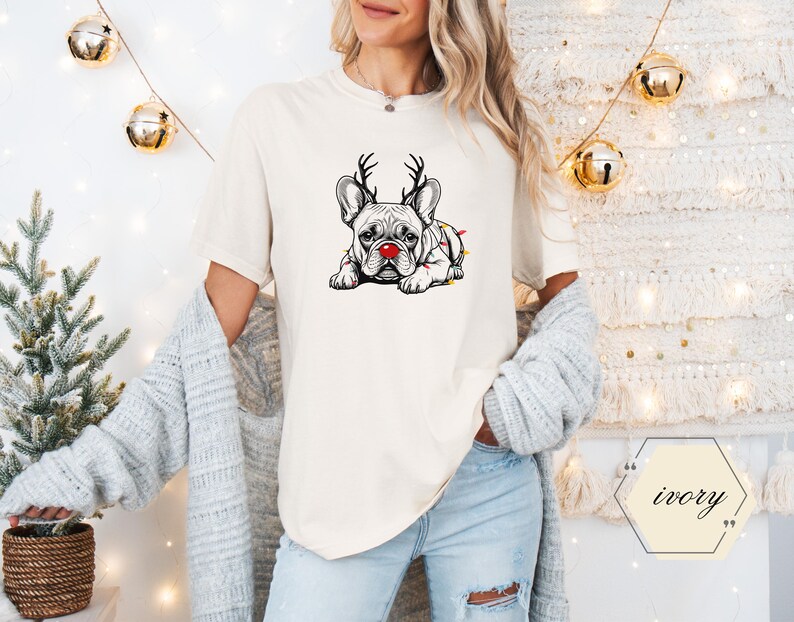 May include: Ivory-colored t-shirt featuring a graphic of a French Bulldog with reindeer antlers and a red nose, wrapped in Christmas lights. The dog is lying down. The shirt is part of a holiday-themed apparel collection.