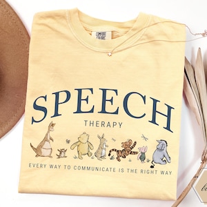 Puede incluir: Una camiseta amarilla clara con las palabras "SPEECH THERAPY" en texto azul marino arqueado. Debajo del texto hay personajes de dibujos animados. La frase "EVERY WAY TO COMMUNICATE IS THE RIGHT WAY" está impresa debajo.
