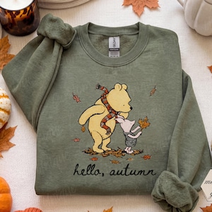 Pode incluir: Uma camisola de gola redonda verde militar com um gráfico do Winnie the Pooh a usar um cachecol às riscas e o Piglet a segurar uma folha. O texto "hello, autumn" está impresso a preto por baixo das personagens, rodeado de folhas.
