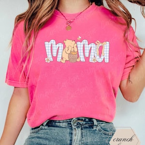 May include: A bright pink t-shirt with the word "Mama" in a blue and white gingham pattern. The design includes a cartoon bear holding a honey pot, a yellow bow, and small flowers. The shirt is a casual, everyday wear item.