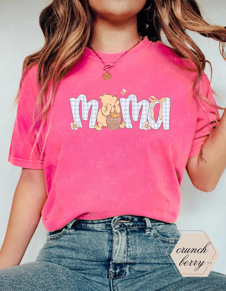 May include: A bright pink t-shirt with the word "Mama" in a blue and white gingham pattern. The design includes a cartoon bear holding a honey pot, a yellow bow, and small flowers. The shirt is a casual, everyday wear item.