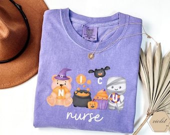 May include: A lilac Comfort Colors t-shirt with Halloween illustrations. The design includes a bear in a witch hat, a bat, a cauldron, a mummy, and a cupcake. The word "nurse" is written in white script below the graphics.