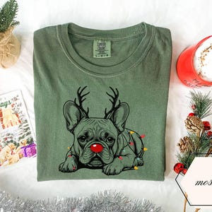 May include: A moss green t-shirt featuring a French Bulldog with reindeer antlers and a red nose, adorned with Christmas lights. The shirt is folded on a white surface, with a holiday card and a red mug nearby.