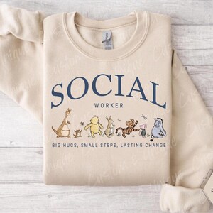 Pu&ograve; includere: Una felpa color sabbia con la scritta "SOCIAL WORKER" in blu navy. Sotto il testo ci sono illustrazioni di personaggi dei cartoni animati. La frase "BIG HUGS, SMALL STEPS, LASTING CHANGE" &egrave; stampata sotto.