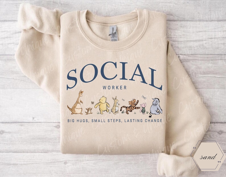 Pu&ograve; includere: Una felpa color sabbia con la scritta "SOCIAL WORKER" in blu navy. Sotto il testo ci sono illustrazioni di personaggi dei cartoni animati. La frase "BIG HUGS, SMALL STEPS, LASTING CHANGE" &egrave; stampata sotto.