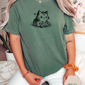 May include: A moss green t-shirt featuring a black and white illustration of a cat playing a video game. The cat is holding a game controller. The shirt is a short-sleeved, crew neck style.