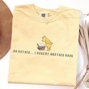 May include: A light yellow t-shirt featuring a cartoon illustration of Winnie the Pooh looking at a stack of books. The text "OH BOTHER... I BOUGHT ANOTHER BOOK" is printed below the illustration. A gold necklace is draped across the shirt.