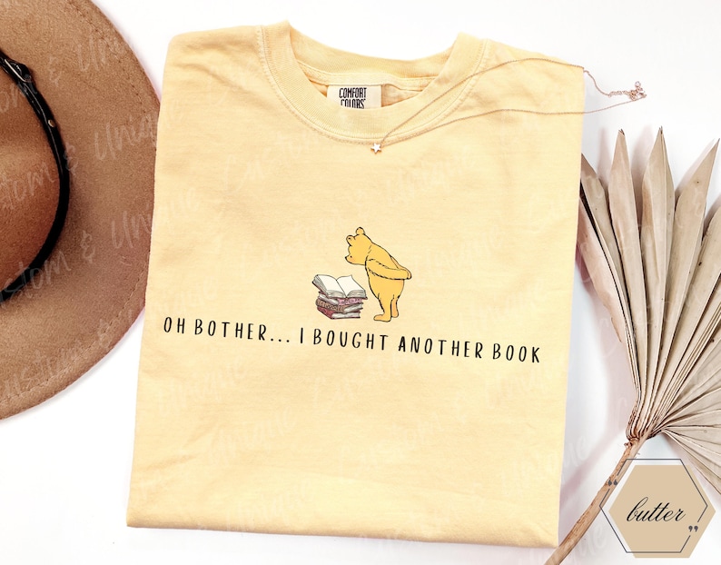 May include: A light yellow t-shirt featuring a cartoon illustration of Winnie the Pooh looking at a stack of books. The text "OH BOTHER... I BOUGHT ANOTHER BOOK" is printed below the illustration. A gold necklace is draped across the shirt.
