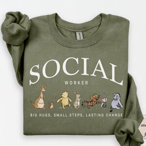 Pu&ograve; includere: Felpa verde oliva con la scritta bianca "SOCIAL WORKER" e personaggi dei cartoni animati. Sotto c'&egrave; la frase "BIG HUGS, SMALL STEPS, LASTING CHANGE". La felpa &egrave; di colore verde militare.