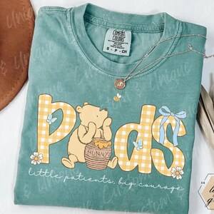 May include: A light green Comfort Colors t-shirt featuring the word "Pods" in a yellow gingham pattern with a Winnie the Pooh graphic. The shirt also has the text "little patients, big courage" and a small bee pendant.