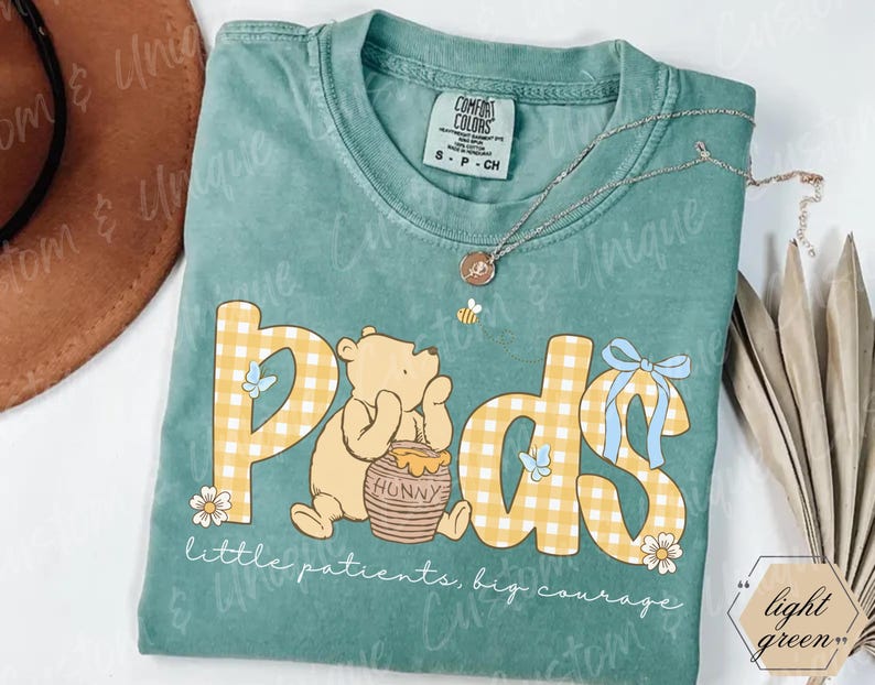 May include: A light green Comfort Colors t-shirt featuring the word "Pods" in a yellow gingham pattern with a Winnie the Pooh graphic. The shirt also has the text "little patients, big courage" and a small bee pendant.