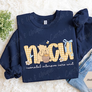 May include: Navy blue sweatshirt with the letters "NICU" in a yellow gingham pattern, featuring a Winnie the Pooh design. The text "neonatal intensive care unit" is below the letters. A blue bow and butterflies are also present.