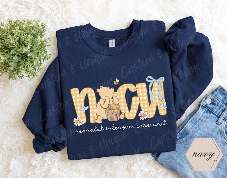 May include: Navy blue sweatshirt with the letters "NICU" in a yellow gingham pattern, featuring a Winnie the Pooh design. The text "neonatal intensive care unit" is below the letters. A blue bow and butterflies are also present.