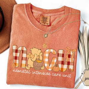 May include: Orange Comfort Colors t-shirt with "NICU" in plaid letters and a Winnie the Pooh graphic. The shirt also features a pumpkin, leaves, and the words "neonatal intensive care unit". A gold necklace and a brown hat are also visible.