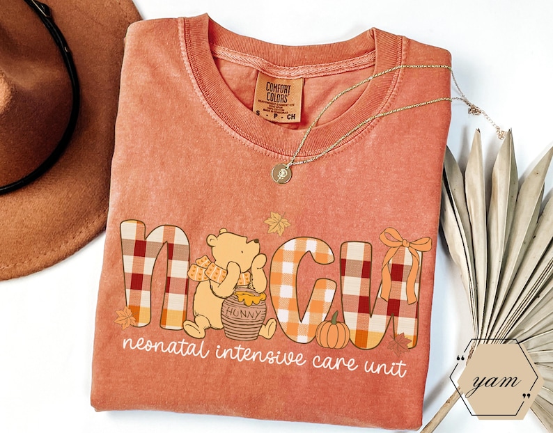 May include: Orange Comfort Colors t-shirt with "NICU" in plaid letters and a Winnie the Pooh graphic. The shirt also features a pumpkin, leaves, and the words "neonatal intensive care unit". A gold necklace and a brown hat are also visible.