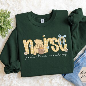 Peut inclure: Sweat-shirt vert for&ecirc;t avec le mot "nurse" en lettres &agrave; carreaux jaunes. Un ours de dessin anim&eacute; tient un pot de miel. Les mots "pediatric oncology" sont &eacute;crits en dessous. Un n&oelig;ud bleu et des fleurs sont &eacute;galement pr&eacute;sents.