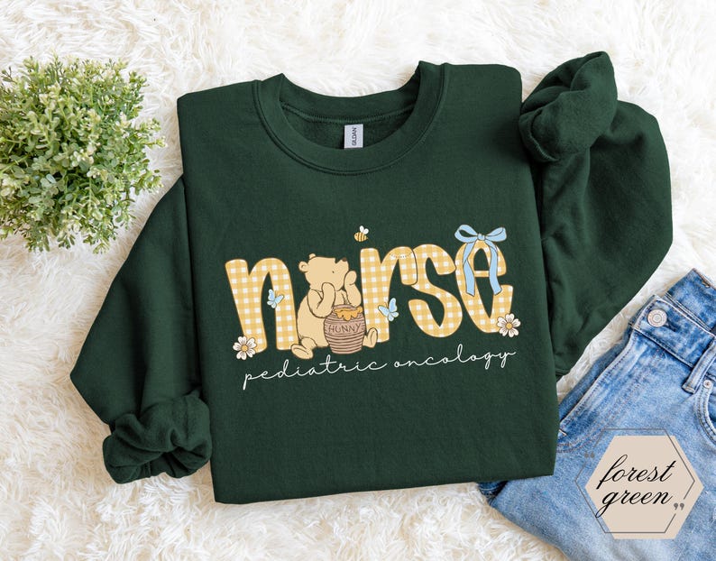 Peut inclure: Sweat-shirt vert for&ecirc;t avec le mot "nurse" en lettres &agrave; carreaux jaunes. Un ours de dessin anim&eacute; tient un pot de miel. Les mots "pediatric oncology" sont &eacute;crits en dessous. Un n&oelig;ud bleu et des fleurs sont &eacute;galement pr&eacute;sents.