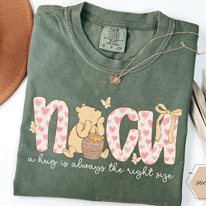 May include: A moss green t-shirt with the word "NCU" in pink hearts and a Winnie the Pooh graphic. The text "a hug is always the right size" is below the graphic. A gold necklace and a brown hat are also in the image.