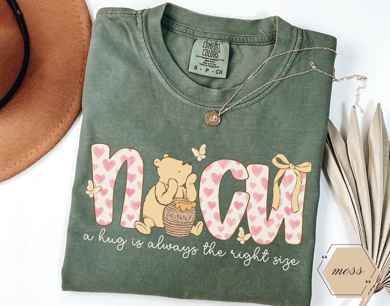 May include: A moss green t-shirt with the word "NCU" in pink hearts and a Winnie the Pooh graphic. The text "a hug is always the right size" is below the graphic. A gold necklace and a brown hat are also in the image.