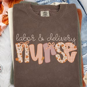 May include: A brown crewneck t-shirt featuring the text labor & delivery nurse in a stylized font. The word nurse is filled with a pattern of orange pumpkins, autumn leaves, and gingham checks, accented with small bows on the letters n and r.