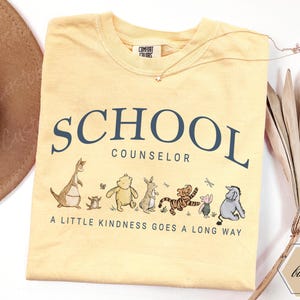May include: A pale yellow crew neck t-shirt featuring the dark blue text SCHOOL COUNSELOR and the phrase A LITTLE KINDNESS GOES A LONG WAY. Below the text is a graphic illustration of Winnie the Pooh characters including Roo, Piglet, Pooh, Rabbit, Tigger, and Eeyore.