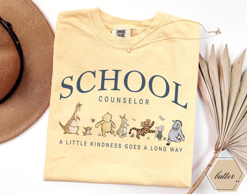 May include: A pale yellow crew neck t-shirt featuring the dark blue text SCHOOL COUNSELOR and the phrase A LITTLE KINDNESS GOES A LONG WAY. Below the text is a graphic illustration of Winnie the Pooh characters including Roo, Piglet, Pooh, Rabbit, Tigger, and Eeyore.