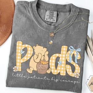 May include: A gray Comfort Colors t-shirt with a Winnie the Pooh design. The design features the word "Pods" in yellow gingham letters, a cartoon bear, and the text "little patients, big courage."