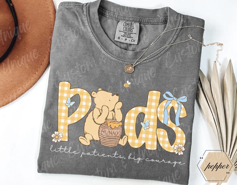 May include: A gray Comfort Colors t-shirt with a Winnie the Pooh design. The design features the word "Pods" in yellow gingham letters, a cartoon bear, and the text "little patients, big courage."