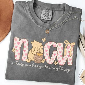 May include: Gray Comfort Colors t-shirt featuring a graphic of Winnie the Pooh with the text "N CU" in pink hearts and the phrase "a hug is always the right size." A gold necklace and a brown hat are also visible.
