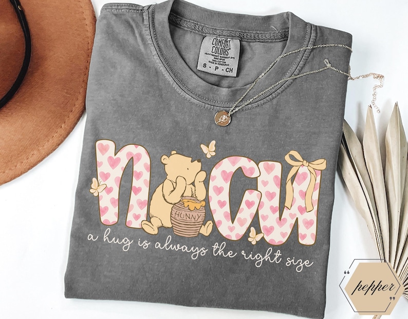 May include: Gray Comfort Colors t-shirt featuring a graphic of Winnie the Pooh with the text "N CU" in pink hearts and the phrase "a hug is always the right size." A gold necklace and a brown hat are also visible.