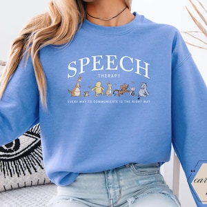 May include: Carolina blue sweatshirt with the words "SPEECH THERAPY" in white arched text. Below the text is a line of cartoon characters. The phrase "EVERY WAY TO COMMUNICATE IS THE RIGHT WAY" is printed below the characters.