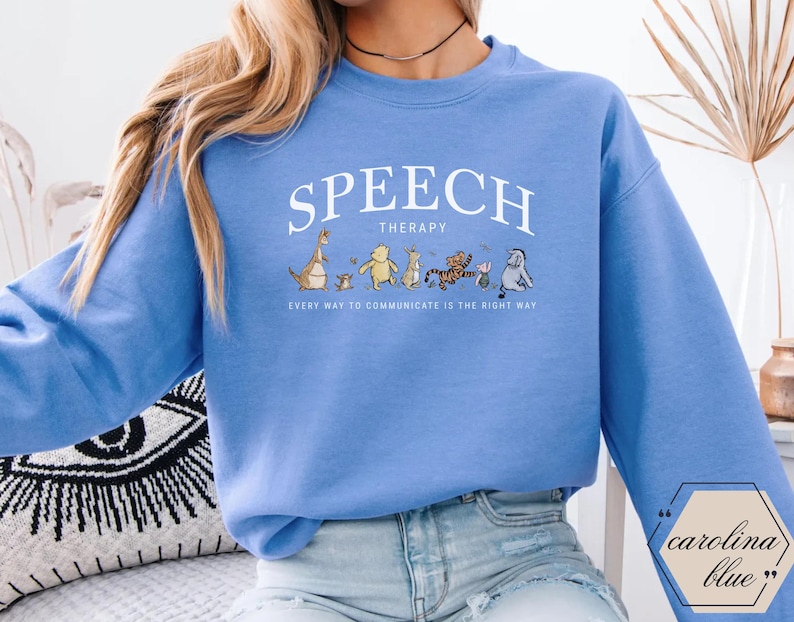 May include: Carolina blue sweatshirt with the words "SPEECH THERAPY" in white arched text. Below the text is a line of cartoon characters. The phrase "EVERY WAY TO COMMUNICATE IS THE RIGHT WAY" is printed below the characters.