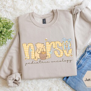 Peut inclure: Un sweat-shirt beige avec le mot "nurse" en lettres &agrave; carreaux jaunes, avec un ours de dessin anim&eacute; et un pot de miel. Le texte "pediatric oncology" est en dessous. Un n&oelig;ud bleu et des fleurs ornent le design.