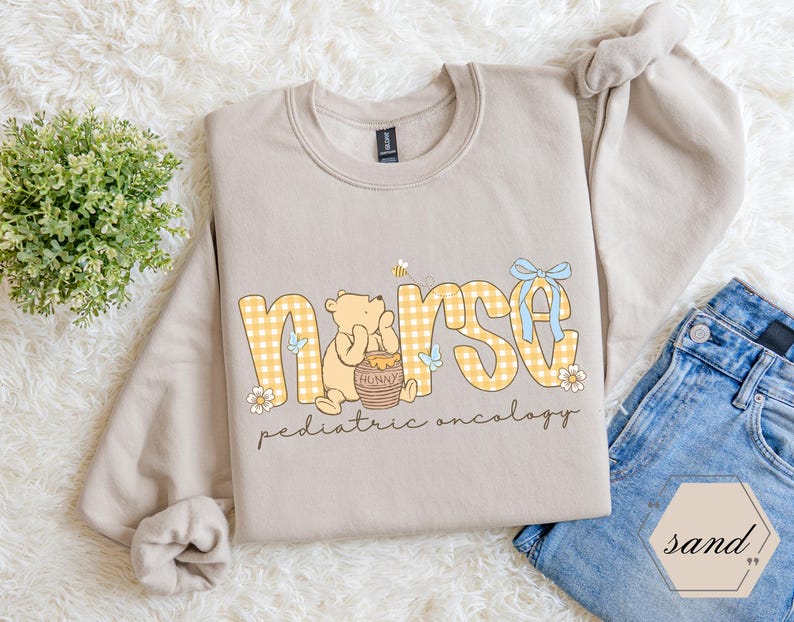 Peut inclure: Un sweat-shirt beige avec le mot "nurse" en lettres &agrave; carreaux jaunes, avec un ours de dessin anim&eacute; et un pot de miel. Le texte "pediatric oncology" est en dessous. Un n&oelig;ud bleu et des fleurs ornent le design.
