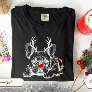 May include: Black t-shirt featuring a white graphic of a French Bulldog wearing reindeer antlers and a red nose, with Christmas lights. The shirt is part of a holiday collection and is a great gift.