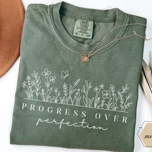 Op de afbeelding: Een mosgroen t-shirt met een wit bloemenontwerp en de tekst "PROGRESS OVER perfection". Een gouden ketting met een hanger ligt op het shirt. Een bruine hoed en een gedroogde plant staan op de achtergrond.
