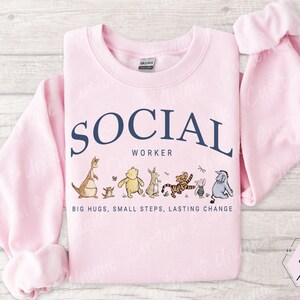 Pu&ograve; includere: Felpa rosa chiaro con la scritta "SOCIAL WORKER" in blu navy, arcuata sopra personaggi dei cartoni animati. La parte inferiore della felpa recita "BIG HUGS, SMALL STEPS, LASTING CHANGE".