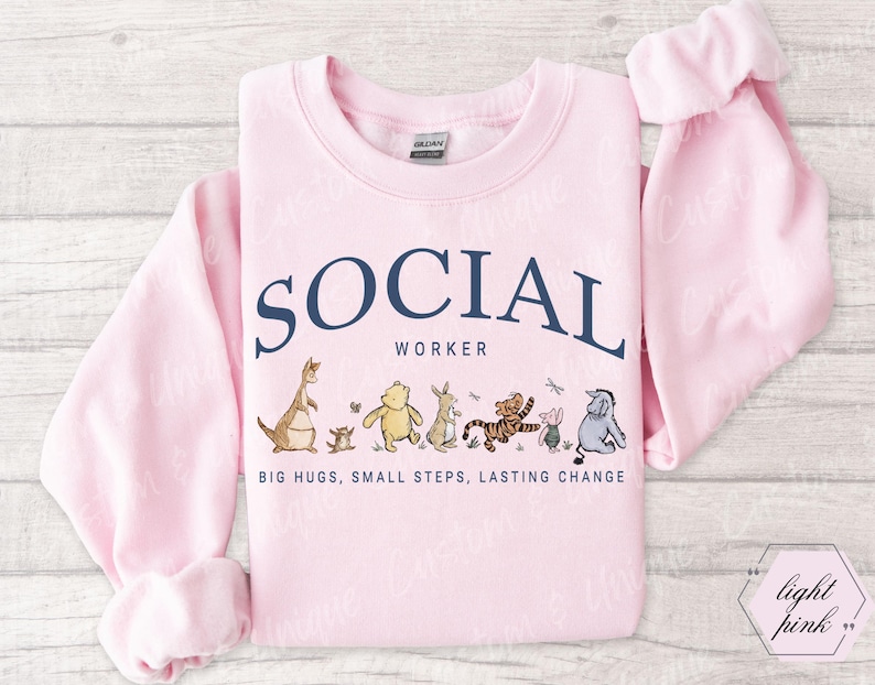 Pu&ograve; includere: Felpa rosa chiaro con la scritta "SOCIAL WORKER" in blu navy, arcuata sopra personaggi dei cartoni animati. La parte inferiore della felpa recita "BIG HUGS, SMALL STEPS, LASTING CHANGE".