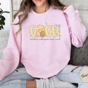May include: Pale pink sweatshirt with "CICU" in yellow gingham, a cartoon bear, and "cardiac intensive care unit". The sweatshirt is made of a soft, comfortable material.