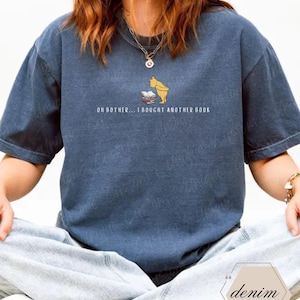 May include: A denim blue t-shirt featuring a graphic of a yellow bear and a stack of books. The text "OH BOTHER... I BOUGHT ANOTHER BOOK" is printed below the graphic. The shirt is a relaxed fit.