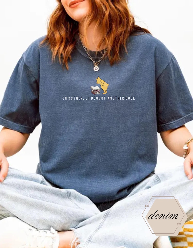 May include: A denim blue t-shirt featuring a graphic of a yellow bear and a stack of books. The text "OH BOTHER... I BOUGHT ANOTHER BOOK" is printed below the graphic. The shirt is a relaxed fit.