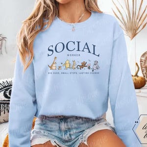 Pu&ograve; includere: Felpa azzurra con la scritta "SOCIAL WORKER" in blu navy. Sotto il testo ci sono personaggi dei cartoni animati. &Egrave; presente anche la scritta "BIG HUGS, SMALL STEPS, LASTING CHANGE".