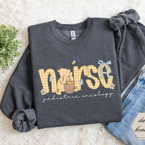Peut inclure: Sweat-shirt gris fonc&eacute; avec le mot "nurse" en vichy jaune, un dessin de Winnie l'ourson et les mots "pediatric oncology". Le sweat-shirt a des manches longues et un col rond.