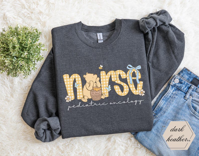 Peut inclure: Sweat-shirt gris fonc&eacute; avec le mot "nurse" en vichy jaune, un dessin de Winnie l'ourson et les mots "pediatric oncology". Le sweat-shirt a des manches longues et un col rond.