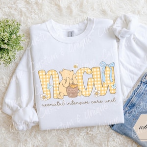May include: White sweatshirt with the letters NICU in yellow gingham print, featuring a cartoon bear holding a honey pot. The text "neonatal intensive care unit" is below the letters. A blue bow and bee are also included.