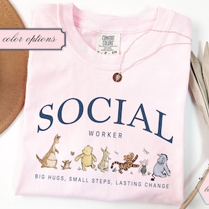 Pode incluir: Uma camiseta rosa claro com as palavras "SOCIAL WORKER" em azul marinho. Abaixo do texto estão personagens de desenhos animados. A camiseta também tem o texto "BIG HUGS, SMALL STEPS, LASTING CHANGE". Um colar de ouro está sobre a camiseta.