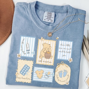 Puede incluir: Una camiseta Comfort Colors azul claro con varios diseños gráficos. La camiseta presenta ilustraciones de Winnie the Pooh, una jeringa, huellas de bebé y el texto "ivf mama". Un collar dorado descansa sobre la camiseta.